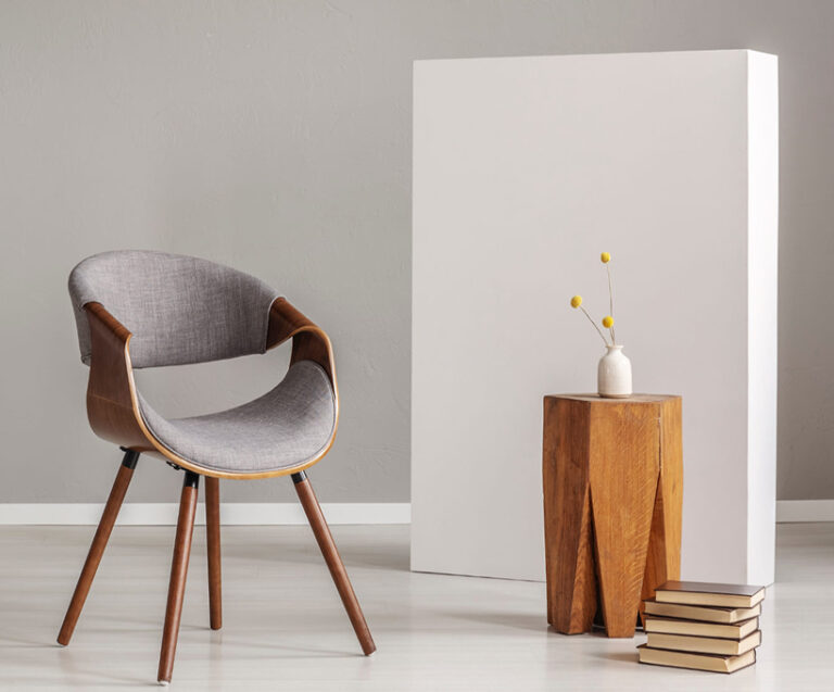 Elegir Muebles Tendencias y tips en kalapa, Interior minimalista con silla moderna de madera y tapizado gris, mesa auxiliar geométrica, jarrón blanco con flores amarillas y panel blanco, ideal para espacios pequeños.