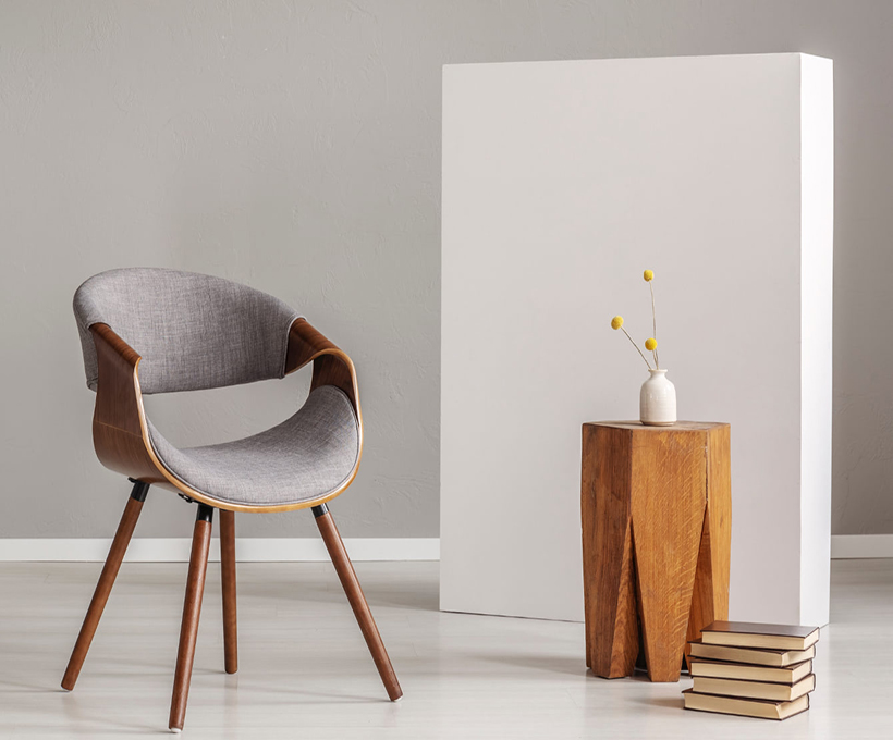 Elegir Muebles Tendencias y tips en kalapa, Interior minimalista con silla moderna de madera y tapizado gris, mesa auxiliar geométrica, jarrón blanco con flores amarillas y panel blanco, ideal para espacios pequeños.